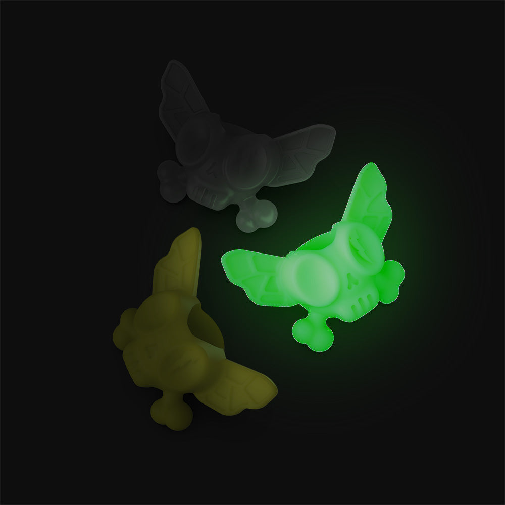 Three silicone beads shaped like flies in yellow, glow in the dark, and translucent gray on a dark background